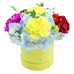 Viewed slightly from above, this compact, joyful bouquet nestles in a round, pastel-yellow gift box that feels both whimsical and elegant. Two plump, ruffled yellow carnations anchor the centre, their layered petals catching light like tiny suns and radiating a warm, uplifting character. Encircling them are generous clusters of powdery periwinkle hydrangeas, their many small florets forming a soft, cloud-like mass that cools and calms the composition while adding tactile fullness. A single deep magenta carnation to the left introduces a vivid note of drama, its saturated tone and sculpted petals creating a lively counterpoint to the airy blues; on the right a velvety crimson bloom brings depth and a touch of formal richness. Sprays of fresh green foliage weave between the flowers, lending slender texture and a natural, garden-fresh scent imagined to the arrangement. The lemon-yellow hatbox is gently patterned with assorted pastel dots - light blue, pale pink and mint green - some with tiny white specks that catch the eye and suggest seasonal cheer. The overall impression is of a carefully curated gift: playful yet refined, ideal for brightening a hospital room, celebrating a milestone, or sending a heartfelt thank-you. As a local florist familiar with Brent Cross and nearby Cricklewood, I picture this design travelling well across neighbourhoods, arriving as a compact burst of colour and scent that reads as both premium and personal.