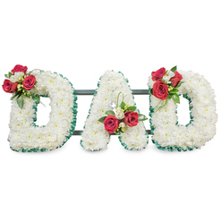 A carefully composed sympathy tribute that spells out DAD in bold, three-dimensional block letters, each letter lovingly covered with densely packed white chrysanthemums that create a plush, serene surface. The small chrysanthemum heads form a uniformly soft carpet of white, their texture almost feathery beneath the fingertips, and they carry a faint, comforting floral aroma. Each letter is neatly outlined with a rich emerald-green ribbed ribbon, offering crisp contrast and a tailored finish that frames the blooms with dignity. Arranged with floral intent, clusters of deep red roses and small white freesia punctuate the design: one arrangement rests on the upper curve of the first D, another nestles inside the central A close to its left leg, and a third mirrors the first on the right D's upper arc. These clusters include glossy green foliage and subtle loops of leaf that lift the composition, adding movement and modern detail without overpowering the solemn simplicity. Slim, dark structural bars are visible behind the letters, supporting the display for a church service or cemetery placement and ensuring each letter remains upright and coherent. The composition is photographed against a clean white backdrop under gentle, diffused light that highlights the contrast between the pure whites and the deep crimson roses. This floral arrangement speaks of quiet remembrance-white chrysanthemums for enduring loyalty and red roses for heartfelt respect-suitable for commemorating a beloved father from homes around Brent Cross, Hendon or Cricklewood.
