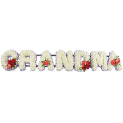 In this image, the loving word GRANDMA is expressed entirely in flowers, each capital letter fashioned into a sturdy block shape and lined up horizontally as though ready to accompany a final farewell at a Brent Cross crematorium or nearby chapel. The letters are densely massed with pure white cushion chrysanthemums, their small, closely set heads creating a soft, uniform texture that looks almost like fine lace when viewed from a distance. A faint halo of lilac or pastel purple ribbon traces the outline of every letter from behind, introducing a subtle, calming tone that gives definition and a sense of quiet grace to the tribute. Vibrant red accents interrupt the white with meaningful touches of colour. On the G, a compact spray of deep red roses is paired with a striking red-and-white calla lily, its variegated petals adding individuality, like a cherished family memory. The first A features a single, heart-shaped red anthurium at its base, shining with a gentle gloss that contrasts beautifully with the soft chrysanthemums. At the centre, the M is decorated with another grouping of rich red roses accompanied by a red anthurium, and the final A is finished with one more anthurium, echoing the earlier blooms so the design feels balanced and complete. The entire word is supported by a discreet dark green stand that keeps the focus firmly on the flowers. Crafted in the manner Brent Cross Flowers often uses for bespoke funeral lettering, this GRANDMA arrangement speaks of gratitude, lifelong devotion, and the warmth of family gatherings, translated into a calm, dignified floral form.