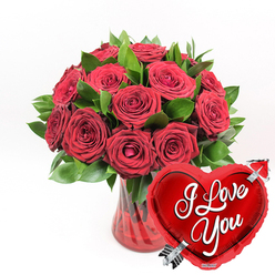 Displayed against a bright white backdrop, a luxuriant bouquet of red roses in a ruby-coloured glass vase takes centre stage, its bold tones and careful styling evoking timeless romance. The arrangement features approximately a dozen or more deep crimson roses gathered tightly together, creating a rounded, abundant silhouette. Each rose is lush and velvety, with petals that curl outward in soft spirals; some flowers are fully open, revealing rich, layered hearts, while others are still partially closed, hinting at emotions that are still unfolding. Threaded among the blooms is an assortment of dark green foliage, its glossy leaves framing the roses and adding volume, while their cool, fresh colour contrasts beautifully with the warmth of the red. The vase itself is a translucent ruby glass, vertically fluted so that light glides along its ridges and intensifies the colour at the base where the rose stems disappear into water. In the right foreground, slightly in front of the bouquet, stands a brilliant heart-shaped foil balloon in bright metallic red. The balloon has a high-shine finish and appears almost three-dimensional, its surface catching reflections from the simple white setting. Across the centre, flowing white script spells out the words "I Love You," and a silver arrow-reminiscent of Cupid's legendary bow-slices diagonally through the heart from lower left to upper right. Together, the roses, vase, and balloon form an unmistakably romantic composition, easily imagined as a gift delivered by Brent Cross Flowers to mark an anniversary near Brent Cross Shopping Centre, a Valentine's Day surprise in Cricklewood, or a thoughtful declaration of love to someone special in nearby Finchley.