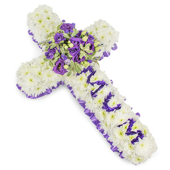 This solemn, thoughtfully arranged floral cross is built from a solid mattress of bright white chrysanthemum blooms, their rounded petals creating a cloud-like surface that reads as both tender and steadfast. The image captures the tribute on a clear white studio background so every detail is visible: a scalloped, deep purple satin ribbon traces the outer edge, its ruffled texture forming a composed border that contrasts with the softness of the chrysanthemums. Running down the upright shaft, the word MUM appears in clearly defined purple letters made from small lisianthus or compact spray chrysanthemums, the letters raised slightly so they stand out against the white field. At the centre a rich focal spray brings the arrangement to life-a compact bouquet of deep violet roses and lighter lavender lisianthus, supported by variegated foliage with pale margins and darker centres, along with tiny green berries that punctuate the cluster and add a subtle, living accent. The palette of snowy white and layered purples suggests a mixture of peace, devotion and dignified sorrow; the design reads as both classic and quietly personal. You can imagine the gentle, powdery scent of chrysanthemums mingling with the sweeter, tea-like perfume of the roses. As an experienced florist in Brent Cross I assemble pieces like this with attention to stability for graveside laying and chapel placement; Brent Cross Flowers can prepare such a tribute for services across Hendon, Cricklewood Broadway and the surrounding neighbourhoods, honouring memory with careful craftsmanship and calm presence.