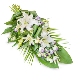 An elegant funeral tribute of white oriental lilies and lisianthus is shown laid diagonally against a stark white background, creating a striking yet soothing contrast. The arrangement takes on a classic teardrop or sheaf shape, with the stems gathered at one end and the flowers and foliage flowing outward in a long, tapering line. Dominating the centre are large, trumpet-shaped lilies in pristine white; some are fully open, their petals smooth and slightly curved at the tips, showcasing pale stamens, while others remain as refined buds, their surfaces satiny and unblemished. Around them, clusters of lisianthus blooms soften the composition, their ruffled white petals often edged with a delicate wash of lavender or lilac, echoing the cool light that often falls across Brent Cross in the early evening. Numerous unopened lisianthus buds punctuate the design, adding rhythm and nuance. Supporting this floral display is a base of varied foliage: broad, dark green leaves provide a quiet, stable background, and a few have been folded and rolled into sculptural twists that add depth without overwhelming the flowers. From one side, elongated palm-style fronds extend outward, their fine lines and gentle arcs lending a sense of graceful motion, like branches moving softly in the breeze near Brent Cross Shopping Centre's landscaped areas. The lighting in the image highlights the natural sheen of the petals and the fresh vitality of the leaves, suggesting a tribute crafted and delivered the same day. This serene spray feels deeply appropriate for moments of farewell, whether placed on a casket, displayed at a local synagogue or church service, or sent as a message of sympathy to a family home in Brent Cross.