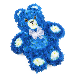 This photograph presents a tender teddy bear-shaped floral tribute resting on a pure white background, the simplicity of the setting drawing all attention to the intricate craftsmanship. The teddy bear's body is formed from a carpet of royal blue flowers, tightly packed so their overlapping petals mimic the softness of fur on a beloved cuddly toy. Creamy white blossoms, with subtle buttery centres, highlight the inner ears, the smooth, rounded snout, and the oval pads on each paw, providing contrast and clarity to the bear's familiar silhouette. The face is completed with two round, dark amber eyes that glint gently, a small black triangular nose, and a delicately curved stitched-style mouth, together giving an expression of calm sweetness and quiet reassurance. Around the neck sits a sheer white organza-style bow, its airy loops and ruffled edges adding a touch of lightness and ceremonial grace. Slightly angled in the frame, the bear feels as though it is peacefully reclining, inviting a sense of soft rest rather than stillness. The fresh petals and crisp colours suggest a piece prepared with great care, ready for same-day delivery across Brent Cross, Golders Green, or Hendon. As a funeral or sympathy tribute, this floral teddy bear would be especially fitting for honouring a baby boy or young child, whether placed by a graveside, in a chapel of rest, or at a family gathering at home, where its gentle blue tones and tender expression could offer comfort and a quiet symbol of enduring love.