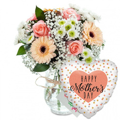 A bright, carefully composed Mother's Day still life is set against a clean white backdrop, every element arranged so that the soft colours and gentle mood can be felt even without seeing the scene. Centrally positioned is a clear glass vase, classic and cylindrical, its base visible through the water and stems. Around the neck, a strand of rustic twine is tied into a neat, casual bow, reminiscent of a florist's finishing touch at a local shop near Brent Cross station. Overflowing from the vase is a rounded bouquet of mixed pastels. Fully opened peach-pink roses sit proudly at the front, their petals curled in generous spirals that suggest both luxury and tenderness. Around them, large peach gerbera daisies spread their layered petals like sunbursts, each bloom anchored by a dark brown centre that adds visual depth and a hint of drama. Weaving throughout the bouquet are light, airy sprigs of white baby's breath, the clusters of tiny blossoms softening every transition between the larger flowers. Small white chrysanthemums, each with a discreet green eye, punctuate the display, creating a rhythm of brightness and gentle contrast. A mix of rich green foliage supports and surrounds the blooms, framing the arrangement and giving it the full, fresh look of something just prepared for same-day delivery across Brent Cross and the nearby residential streets. Slightly hidden behind the flowers rests a small white greeting card, only a sliver of its colourful writing visible, hinting at a personal note of love and thanks. In front of the vase and shifted a little to the right, a large heart-shaped foil balloon adds a playful, celebratory accent. The balloon is white with scattered metallic gold and silver confetti dots that catch the light, creating a subtle sparkle. At its centre a peach-coloured heart encloses the black lettering "HAPPY Mother's DAY," with "Mother's" written in flowing script that feels affectionate and sincere. The overall impression is of a warm, joyous gift moment, like a family surprise waiting in a living room near Brent Cross after a day out together.