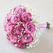 Poised gracefully atop a light, subtly textured beige fabric, this bridal bouquet radiates contemporary elegance rooted in Brent Cross's bustling community. At the heart of the arrangement are ten to twelve roses, their petals carefully layered in a seamless gradient from pale lavender-pink at the core to vibrant fuchsia along the fringes-a spectrum of hues reminiscent of Brent Cross's vibrant spring gardens. Swirling among the roses, clouds of dainty white baby's breath provide a delicate, airy counterpoint that elevates the bouquet's romantic mood. Each bloom, perfect in form and freshness, is nestled tightly for a modern domed silhouette that fits comfortably in the hand. The stems extend below a wrap of blush pink satin ribbon, finished with a slender spiral of luminous white pearls that add an opulent, classic finish. Soft, natural light bathes the bouquet, bringing alive every detail-the velvety rose contours, the sparkling gypsophila, and the gentle sheen of ribbon and pearls. The background's neutral simplicity ensures that all the focus rests on the colours and textures of the flowers, much like an understated yet striking center table at a Brent Cross wedding or family celebration. This bouquet embodies love and celebration, perfect for commemorating unions, expressing affection, or simply marking life's joyful moments with style and artistry from Brent Cross Flowers.