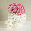 A softly lit display features a compact dome of pink roses cradled in an artistic white orb, creating a wedding arrangement that radiates calm sophistication, much like a peaceful afternoon in the quieter corners of Brent Cross. The roses themselves are meticulously placed, their tightly layered petals forming smooth, rounded heads that touch and overlap in a continuous wave of colour. Each flower glows with subtle gradients, transitioning from gentle blush at the core to a deeper, more saturated pink at the edges, suggesting the care of an experienced florist's hand. Around this central cluster of roses, a ring of white baby's breath rises like delicate mist. The tiny gypsophila blooms add volume without weight, introducing a soft, cloud-like frame that contrasts beautifully with the boldness of the pink petals. The arrangement is anchored in a contemporary white globe, woven from slender strands that curve and intersect to form an open, basket-like shell. This airy structure allows the stems and interior shadows to show through, offering a sense of depth and visual intrigue. Beneath the orb, two minimal, geometric ornaments sit quietly on the tabletop, reflecting the same modern design language and ensuring the eye moves gracefully around the scene. The neutral background keeps the attention firmly on the interplay of pink and white, evoking a sense of serenity and refined joy. Perfect for wedding breakfasts in Brent Cross hotels or intimate family receptions at home, this composition speaks to couples who appreciate both classic romance and clean, modern style in their wedding flowers.