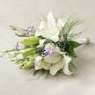 An elegant, hand-tied bouquet lies horizontally on a neutral, softly woven surface, echoing the refined ambience of Brent Cross events. The arrangement is dominated by spectacular Oriental lilies, their snowy white, recurved petals delicately sprinkled with freckles and adorned by vibrant yellow anthers, exuding purity and celebratory spirit. Scattered throughout are clusters of white lisianthus edged in rich purple, their semi-open blooms and closed buds providing dynamic interplay of colour and texture, reminiscent of Brent Cross's diverse floral markets. Slender purple spikes-likely Veronica-project upward, infusing the design with depth and a sense of lively movement. Cloud-like clusters of baby's breath float gently between larger flowers, adding softness while harmonising the bouquet. A lush frame of green foliage, from broad-leaf bases to cascading, fern-like stems, brings freshness and fullness, echoing the verdant spaces of North London. Tightly bound stems finish with a simple white ribbon, showcasing the meticulous style of local Brent Cross florists. Bathed in gentle, even light, every detail is rendered clear and inviting, making this bouquet an ideal bridal choice or thoughtful gesture for celebrations near Brent Cross Shopping Centre.