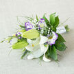 Spread along a softly woven, light beige fabric that resembles natural linen, this bouquet lies at ease as if just completed in the Brent Cross Flowers studio, ready to be carried down the aisle at a local ceremony. The dominant blooms are white Oriental lilies, some fully open with their broad, gently arching petals, others firm and upright as long green buds. On the open lilies, slender stamens extend from the heart of each flower, their orange-yellow tips holding a dusting of dark pollen that contrasts beautifully with the creamy white petals, which are lightly speckled on the inside. Interwoven with the lilies are lisianthus in a romantic mix of white and purple; the petals start as clean white at the centre and gradually deepen to lilac and rich violet at the edges, their ruffled forms suggesting hand-painted china or watercolour touches. Several lisianthus buds, tightly closed and gently tinged with purple, contribute an air of youthful freshness. Scattered throughout the arrangement, delicate sprays of baby's breath float like tiny white stars, filling spaces and lightening the overall composition. A generous selection of greenery provides structure and depth: broad, glossy dark green leaves, feather-light fronds, and thin, reed-like stems combine to give the bouquet a layered, textural feel, reminiscent of the varied greens around nearby Clitterhouse Playing Fields. The stems are drawn together in a neat bundle and wrapped with a white satin ribbon, which lies smooth and lustrous against the stalks, accented with minute pearl-like beads that catch the light in a subtle, celebratory sparkle. This bouquet feels both intimate and refined, ideal for weddings and special occasions around Brent Cross, evoking calm joy, loyalty, and a graceful sense of anticipation.