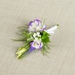 A finely detailed boutonniere lies against a softly textured, light beige linen-style fabric, the neutral background allowing every petal and leaf to stand out with quiet elegance. At its heart are two open lisianthus blooms, their petals a creamy white with delicately ruffled edges brushed in a vivid, royal purple that ripples like ink along fine paper. Nestled closely around them are tiny clusters of crisp white filler flowers, reminiscent of baby's breath, giving the piece a cloud-like softness. A slim vertical spire of deep purple muscari buds rises beside the lisianthus, each tiny bell-shaped flower tightly packed and rich in colour, adding a graceful line and extra depth to the design. Pale green, unopened buds peek out from among the blooms, suggesting movement and freshness, as though the flowers were gathered only moments ago from a garden near Brent Cross. Fine, feathery green foliage, similar to asparagus fern, fans gently around the flowers, its airy texture framing the composition and lending a natural, slightly wild charm that still feels perfectly tailored for a formal lapel. The exposed stems are neatly aligned and wrapped in a smooth band of white satin ribbon, finished with a small, precise bow that hints at the care taken by an experienced florist preparing it for a groom or prom guest. The lighting is soft and even, with no harsh shadows, giving the entire boutonniere a calm, polished feel, ideal for a wedding at a Brent Cross venue or a special celebration in nearby North London neighbourhoods.
