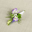 This boutonniere offers a small, carefully composed story of spring - two ruffled lisianthus flowers, white with a cool violet edge, stand at the heart of the design like a tender promise. Around them, tiny clusters of baby's breath float like miniature clouds, providing a whisper-light backdrop that softens the silhouette. Below, the vertical spikes of grape hyacinth introduce concentrated blue-violet colour, their bell-shaped florets packed tightly to create a lively visual contrast against the lisianthus' open faces. Feathery fronds of fern sweep outwards, adding an organic, woodland texture that balances the composition and gives it a graceful, airy outline. Pale buds and slender stems extend with a gentle rhythm, suggesting movement and the fresh energy of early spring. The stems are gathered and secured with a white satin ribbon, knotted with care so the piece can be pinned neatly to a lapel or held briefly before being attached during a wedding, anniversary, or portrait session. Arranged on neutral linen under soft daylight, the cool palette of whites, violets, and blues reads as composed yet intimate. As a local florist familiar with Brent Cross and nearby Cricklewood, I picture this boutonniere paired with crisp tailoring for a ceremony at the shopping centre chapel or an understated family celebration; Brent Cross Flowers offers local collection and delivery so this small, fragrant detail arrives ready to be worn and remembered.