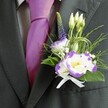 A close-up photograph shows the upper torso of a man dressed for a wedding or smart Brent Cross celebration, the focus resting on a carefully tailored dark charcoal grey suit jacket and an exquisite buttonhole. The fabric of the jacket looks softly textured yet structured, its lapel smooth and clean, framing the floral detail with quiet confidence. Beneath the jacket, a pale grey checkered shirt adds a refined pattern, while a lustrous magenta silk tie, perfectly knotted, brings a vivid flash of colour that immediately suggests festivity and pride. Pinned to the left lapel is a detailed boutonniere built around white lisianthus blooms edged in deep purple, their ruffled petals gathered like miniature watercolor clouds. At the centre of each flower sits a gentle yellow eye, drawing attention and echoing the warm mood of the day. Around these main flowers cluster slender white buds not yet open, tiny white filler flowers similar to baby's breath, and a single vertical spike of dark indigo veronica, which adds height and elegance. Fine, feathery greenery mingles with broader emerald leaves and looping blades of decorative grass, giving the arrangement movement and a fresh, garden-picked feel, as if it could have come from a morning walk through nearby Brent Cross parks. A petite white satin bow at the base secures the stems and finishes the look with a timeless, traditional touch. The overall impression is of poised, modern romance: crisp tailoring, rich colour, and lovingly crafted flowers all working together to capture the excitement of a wedding day or formal event in North West London.