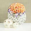 This composition feels like a quiet sunrise on the table: a compact, domed cluster of some two to three dozen apricot and soft-peach roses, each bloom at a slightly different stage so the bouquet carries movement from tight buds to fully opened cups. Nestled at the perimeter, white Lisianthus edged in a deep violet lend a soft, painterly contrast that lifts and defines the shape, while delicate sprigs of Gypsophila are scattered throughout to introduce lightness and a vintage, romantic air. The blooms are cradled in a sculptural white woven vessel - an airy, reed-like orb with an open lattice - which reads contemporary yet handcrafted and invites a hand to trace its texture. To the left on the spotless surface sit two decorative rattan or bamboo spheres, one larger and one smaller, their pale wood strands woven into spherical cages that echo the basket's geometry and enhance the arrangement's natural aesthetic. The background is a subtle linen-beige, gently out of focus, allowing the warm tones of the roses and the violet accents to remain the visual centre. You can almost imagine the scent: the honeyed warmth of apricot roses balanced by the green-fresh lift of lisianthus and the faint sweetness of baby's breath. Perfect as a table centre for an intimate Brent Cross gathering or as a thoughtful gift for anniversaries and new homes in nearby Cricklewood and Hendon. As a local florist, I would prepare this for careful local delivery across Brent Cross and adjoining neighbourhoods, ensuring the weave and blooms arrive intact and fragrant.
