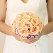 A close-up, chest-level photograph shows a bride standing serenely, her torso framed against a soft, pale background that feels like gentle daylight filtering into a Brent Cross townhouse or registry office. She wears a classic white wedding gown with a fitted lace bodice; the lace is delicate and slightly sheer at the shoulders, forming a modest V-neckline that draws the eye naturally down to the bouquet she cradles. Her hands, resting just in front of her waist, are neatly manicured in a light, neutral polish, with a hint of shimmer catching the light on one thumb, echoing the quiet sparkle of a special day. Nestled in her grasp, the round bridal bouquet dominates the image. At its heart sit dozens of closely packed roses in soft peach, blush pink, and light apricot tones, their petals tightly spiralled and satin-smooth. They create a domed cluster, each bloom gently cupping the next, suggesting warmth, tenderness, and romantic abundance. Beneath this cloud of roses, a rich ring of deep purple flowers edged in creamy white-likely lisianthus-forms a vivid collar, adding depth and a slightly unexpected twist of colour, much like a stroll from Brent Cross Shopping Centre into the quieter surrounding streets. Tiny sprays of airy white baby's breath peek between the petals at the base, softening the transition from rose to purple and giving the bouquet a lace-like halo that mirrors the gown's detailing. The overall effect is refined yet heartfelt, capturing the still, anticipatory moment before the bride walks down the aisle, the bouquet poised as both accessory and symbol of the promises to come in this North West London celebration.