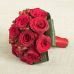 Resting gently on a light beige, linen-like fabric that looks warm and inviting, this compact bouquet of red roses exudes a sense of classic romance and meticulous design. The flowers are arranged in a tight, rounded cluster, each bloom pressed close to the next so that their velvety petals form an almost seamless surface of crimson and burgundy. Looking closely, you can imagine the softness of each petal, the edges slightly ruffled, the centres tightly swirled, hinting at freshly opened roses in their prime. Between the blooms, clusters of glossy red hypericum berries punctuate the arrangement, adding delightful dots of light and texture that catch the eye and enliven the design. Around the base, broad dark green leaves are folded and arranged with precision, forming a polished collar that not only frames the roses but adds a structured, architectural quality. The stems are gathered into a clean handle, wrapped in a bold red satin ribbon that gleams subtly, reinforcing the bouquet's rich colour palette. The composition is photographed from above at a gentle angle, letting the viewer appreciate both the rounded overall shape and the intricate details. It is easy to imagine this bouquet being carried down an aisle in Brent Cross, perhaps on the way to a ceremony near Clitterhouse Playing Fields or presented during an intimate dinner at home after a day shopping at the mall. The mood is undeniably romantic, perfect for marking milestones and special celebrations with flowers that feel both luxurious and deeply personal.