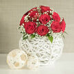 At the heart of the image stands an elegant arrangement of red roses, their petals a rich, saturated crimson that spirals inward with velvety precision. The roses are grouped so closely that their heads form a neat, rounded mound, suggesting a dozen or so carefully selected stems chosen at the same stage of bloom. They are framed by a delicate collar of white baby's breath, each stem branching into clusters of tiny, snow-like blossoms that create a soft, clouded edge to the bouquet. Subtle touches of dark green leaves emerge from beneath the flowers, adding depth without stealing focus from the interplay between red and white. This crowned bouquet is nestled into a contemporary white sphere-shaped vase, its surface made from fine rods woven into a crisscross pattern that recalls hand-crafted wicker yet feels sleek and modern.

The vase rests on a pristine white table, where the open structure allows pockets of light to fall through, sketching faint patterns that echo the lattice design. Two decorative balls sit to one side, one larger, one smaller, made from thinner, cream-toned strands that look like pale rattan or dried vine. Their neutral shade harmonises with the vase and tabletop while providing a gentle contrast in texture, like stylish accents you might spot in a Brent Cross flat overlooking the North Circular or a newly renovated home near Clitterhouse Playing Fields. The background is a soft, neutral wall in beige or light grey, free of clutter so that all attention centres on the flowers and their sculptural surroundings. The atmosphere is calm, romantic, and refined, perfectly suited to a wedding table arrangement, proposal dinner, or heartfelt gift, whether placed in a restaurant near Brent Cross or delivered as a surprise to a loved one at home.