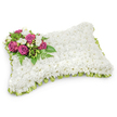 A tender, pillow-shaped funeral tribute, carefully assembled from hundreds of small, pristine white chrysanthemums that form a densely packed, fabric-like surface across a rectangular base with softly rounded corners. The arrangement reads as a gentle cushion of blooms - the chrysanthemums' tight petals and faint herbal scent suggesting quiet purity and peaceful rest - while a single flourish of colour gathers in the top-left corner as a visual and emotional focal point. There, rich magenta and deep fuchsia roses open gradually, their velvety petals catching a soft studio light that gives them a satin sheen; between the roses nestle sprays of delicate white freesias whose subtle fragrance and tapered buds add air and contrast. Lush green foliage, including slender fern fronds and glossy leaves, fans behind the spray to lift the composition and reinforce a living, natural underpinning. Around the entire edge a pleated border of pale green satin ribbon peeks out from beneath the flowers, its gentle sheen providing a refined frame and a whisper of fresh colour against the purity of the white surface. The pillow sits against a clean white background with a faint, natural shadow beneath, allowing every texture-from the chrysanthemum's softness to the rose's richer grain-to be read clearly. This classic sympathy design, familiar at funerals and graveside services across Brent Cross and nearby Cricklewood, is meant to convey comfort, repose and enduring remembrance when words feel inadequate - a quiet floral embrace for those who come to say goodbye.