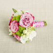 On a softly textured, light beige cloth that looks like natural linen spread across a table in a Brent Cross townhouse, a petite bouquet lies ready to be pinned or placed in waiting hands. The arrangement is compact but full of character, built around two lush roses whose petals show a gentle gradient from tender blush pink at the tightly cupped centres to deeper, saturated fuchsia and magenta at the edges. The roses feel velvety and alive, as though they still hold the morning dew from a North London market. Threaded between them, lime-green button chrysanthemums cluster in neat, globe-shaped forms, each head covered in tiny petals that give a crisp, almost playful texture and a sparkling contrast to the romantic roses. Scattered between these focal flowers are small white blooms, some fully open with slender petals and soft yellow-green cores, others still curled as creamy buds that promise to unfold. Slender green stems and a hint of foliage peek from beneath, adding just enough structure and natural rhythm. The stems are carefully bound together in a short, comfortable handle, wrapped in a rich fuchsia satin ribbon that echoes the rose tones. Around the ribbon, a fine strand of pearl-like beads spirals delicately, catching the light and bringing a touch of bridal elegance. The whole piece feels perfect for a wedding boutonnière, a wrist corsage, or a small bridesmaid bouquet for a celebration in and around Brent Cross, capturing the mood of quiet joy, new beginnings, and considered floral artistry.