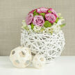 Set atop a sleek white tabletop, polished enough to reflect subtle glimmers of colour, this floral arrangement has the poised composure of a wedding centerpiece in a Brent Cross restaurant or boutique hotel. The focal point is a sculptural sphere of white-painted branches, carefully intertwined to form an open, woven vase that feels at once modern and gently rustic. From this airy cradle emerges a perfect floral dome, rounded and full, with every bloom facing outward in a harmonious display. Pastel pink roses unfold generously, some with their outer petals touched by a slightly richer pink, their velvety surfaces catching the soft, ambient light. Nestled between them, compact lime-green button chrysanthemums form tight, cheerful clusters, their fresh, grassy tone brightening the arrangement and giving it a contemporary twist that would suit a stylish event near Brent Cross Shopping Centre. The base of the bouquet is softened by a ring of tiny white blossoms-possibly sprigs of lisianthus or a fine stock-whose small, starry flowers and pale centers appear to cascade gently over the rim of the vase. To the left, two decorative balls made from pale, interwoven wood strips rest quietly on the tabletop, one larger, one smaller, their open framework reflecting the vase's design and adding visual balance. The background is a blurred, warm neutral wall with a subtle woven or linen-like texture that adds depth without distraction. Altogether, the scene conveys a sense of calm, freshness, and refined celebration, ideal for a wedding breakfast, anniversary dinner, or engagement toast held in or around Brent Cross, where tasteful simplicity meets thoughtful design.