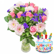 A vibrant, celebratory bouquet rests against a crisp white background, the kind of arrangement you would imagine carried into a small Brent Cross flat to mark a birthday morning. At the centre, a generous cluster of soft pink roses unfurls - petals layered like silk, some a pale blush and others a touch deeper to give the bouquet a gentle, romantic warmth. Around them sit large, pale-pink Gerbera daisies whose broad, open faces and buttery yellow centers read like smiles; their petals range toward almost-white at the tips, catching the light. Scattered through the composition are tight clusters of vivid purple filler flowers, likely statice or limonium, whose papery texture and concentrated colour add depth and a lively contrast to the pinks. Small white spray blooms nestle in the shadows, lending delicate highlights. Glossy deep-green leaves form a lush frame, their smooth surfaces and varied shapes giving the arrangement structure and a fresh, garden-picked feel. The stems are visible through a clear glass vase with a slightly wider base; water and green stalks show at the bottom, reinforcing freshness and the florist's careful conditioning. To the lower right, a cheerful cake-shaped Mylar 'Happy Birthday' balloon leans against the bouquet: predominantly white like frosted cake, trimmed with a wavy blue icing border and scattered with colorful sprinkles in red, yellow, blue and green. Four bright candles - purple, red, orange and blue - stand on the cake illustration with tiny yellow flame tips, and the phrase Happy Birthday arcs across the front in playful, multi-coloured letters. The small brand name KALEIDOSCOPE is tucked at the balloon's base. Soft, natural light suggests a gentle fragrance of roses and fresh greens; it feels like a thoughtful, joyful gift ready for collection or local delivery across Brent Cross.