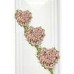 A trio of heart-shaped floral arrangements appears to float down a white wall, forming a vertical cascade that feels both elegant and deeply personal. Each heart is traced in living green, with delicate vines and ivy-like stems intertwining to create soft, natural outlines. These leafy borders are neither rigid nor perfectly symmetrical; instead, they curve and twist as if gently shaped by hand, echoing the kind of bespoke craftsmanship you might expect from a local Brent Cross florist. Within each heart, a dense concentration of petite roses fills every space, their petals unfolding in layers of dusty lavender, mauve, and pale pink. The blooms are arranged so closely that they create a plush, textured surface, like a cushion of petals into which light quietly settles. The top heart sits slightly to the left, the second follows below with a subtle tilt to the right, and the third continues the same progression, each heart connected to the next with narrow threads of greenery, giving the impression that all three are part of one living garland. Soft, even lighting falls across the tributes, enhancing the pastel tones and the velvety quality of the roses without creating harsh shadows. The background is completely clean and uncluttered, focusing attention solely on the shapes and colours of the hearts. This composition is especially suited to funerals, cremation services, or memorial gatherings in and around Brent Cross, where families wish to express enduring love and tender remembrance. The triple-heart motif suggests connection, shared memory, and the continuing presence of a loved one in the hearts of those left behind.
