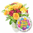 A bright, comforting Get Well Soon gift set framed against a clean white background: on the left a lush bouquet blooms with soft, warm tones and on the right a shiny round foil balloon lifts the sentiment. The bouquet reads as a small celebration of recovery - several large orange roses with gently unfurling petals form the heart of the arrangement, their warm apricot tones softened by the pale edges of each blossom. Sun-bright yellow gerbera daisies punctuate the composition, their flat faces and dark centers like small suns, while deep magenta or burgundy carnations add ruffled texture and visual weight between the roses. Little sprays of white alstroemeria (Peruvian lilies) peek through, bringing delicate flecks of light across the mass of color, and is finished with abundant fresh green foliage whose glossy leaves cradle each bloom. The flowers sit in a clear glass vase with a subtle frosted or textured white pattern that gives the presentation a quietly elegant base; you can imagine the cool clarity of the water and the tidy stems inside. To the right, overlapping slightly, a metallic silver Mylar balloon provides a playful backdrop: a compact cluster of cartoon balloons in blue, purple, pink, yellow and green decorates the center, some striped or polka-dotted, circled by tiny starbursts along the white rim. Bold yellow-and-blue letters declare Get Well Soon, and a tiny KALEIDOSCOPE logo is visible near the balloon's lower edge. Bathed in soft daylight, this arrangement radiates warmth and hopeful cheer - a thoughtfully composed hospital or home delivery choice for someone in Brent Cross, Cricklewood, or nearby Hendon seeking a lift in spirits.