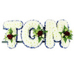 This photograph captures a carefully crafted floral name tribute for TOM, a design often ordered for funerals and memorials in the Brent Cross area when families want to honour a loved one personally. The three letters are shaped as bold block capitals, mounted in a row on a discreet dark frame that offers strong but almost invisible support. Every visible surface of the T, O, and M is densely covered with immaculate white chrysanthemums, placed so close together that they form a thick, cushion-like layer of soft petals, suggesting purity and peace. Tracing the outside edge of each letter is a royal blue ribbon, crisply pleated and pinned to create a clear outline that makes the name stand out against the plain white background, much as it would against the interior of a chapel or crematorium near Brent Cross or Cricklewood. At thoughtfully chosen points across the letters, small yet striking sprays of mixed flowers add colour and character. Deep, velvety red roses act as the focal blooms, accompanied by smaller cream and white spray roses and delicate, slender white freesia-like flowers that introduce a hint of lightness and fragrance. Around these clusters, fresh green foliage-feathery ferns and assorted leaves-spills gently over the chrysanthemums, bringing depth, contrast, and a touch of natural movement, reminiscent of local parks and gardens. The overall impression is one of quiet dignity and heartfelt remembrance: a bespoke funeral tribute that spells out Tom's name with clarity and tenderness, suitable for delivery to churches, chapels of rest, and crematoria throughout Brent Cross and the surrounding North London community.