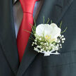 The image captures a close-up of a wedding boutonnière sitting neatly in the pocket of a dark, finely tailored suit, the charcoal fabric forming a strong, structured backdrop. Rising from the pocket, a single white orchid unfurls, its smooth petals spreading with serene grace and a gentle, luminous sheen, like soft light reflecting off winter clouds above Brent Cross. The orchid's centre holds a hint of golden cream, adding a quiet glow that invites a longer look. Surrounding the bloom, airy sprays of baby's breath create a delicate, cloud-like frame, each tiny white flower contributing to an impression of purity and softness. Slim shoots of fresh greenery thread through the arrangement, offering subtle depth and movement, as if a breath of garden air has been tucked into the formal suit. The stems are gathered at the base with a slender white ribbon, its satin folds precise and tidy, revealing the hand of an experienced florist. Just behind the boutonnière, a vivid red silk tie descends in a bold streak of colour, its sheen and intensity heightening the contrast with the pale, gentle tones of the orchid and gypsophila. The overall design feels both classic and contemporary, ideal for a groom arriving at a ceremony near Brent Cross tube station or a reception venue tucked close to the North Circular. It carries the mood of a joyful union: quiet anticipation, love worn close to the heart, and flowers chosen with care to reflect the significance of the day and the personality of the wearer.