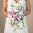 Soft late morning light filters through this close-up scene, where a bride in Brent Cross stands mid-frame, her gentle arms wrapped around an exquisitely composed cascading bridal bouquet. She wears a strapless white gown, trimmed in lace that subtly catches the light, providing an elegant contrast to the central floral arrangement. Domineering the bouquet are stately white phalaenopsis orchids-petals gleaming and centres touched with yellow-characterizing the modern chic favoured at nearby Brent Cross events. Around them, plush garden roses in a pastel blush pink lend timeless romance, while clusters of pale lilac and lavender stock and lisianthus introduce scattered pops of colour and nuanced textures. Baby's breath fills the spaces like a mist, and wisps of green foliage offer natural balance without competing for attention. The bouquet arches elegantly, descending in soft waves to mirror the curves of the dress. Altogether, this scene speaks of quiet sophistication and careful craftsmanship, the work of florists passionate about creating unforgettable bridal bouquets for the Brent Cross community, echoing the joyful energy of weddings celebrated from Golders Green to Hendon.