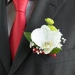 A detailed close-up focuses on a single wedding boutonniere pinned neatly to the lapel of a dark charcoal grey suit jacket, the fine wool fabric showing a subtle woven texture in the soft, even light. At the heart of the design sits a large white Phalaenopsis orchid, its broad, velvety petals opening like wings, with a delicate yellow and white lip drawing the eye to the centre. Tucked gently around the orchid, tiny stars of white baby's breath form a cloud of softness, while two small, button-like lime-green chrysanthemum blooms add a bright, playful contrast above. A few rich green leaves frame the flowers, their smooth surfaces catching a gentle sheen that suggests freshness and careful conditioning. The stem of the boutonniere is wrapped with a narrow deep red satin ribbon, its sheen echoing the bold, solid red tie that runs vertically to the left against a crisp, light grey and white small-checked dress shirt. The lighting is natural and flattering, enhancing every curve of petal and line of stitching on the visible breast pocket beneath. The layout suggests a groom or groomsman standing ready for a ceremony in Brent Cross, perhaps near the busy shopping centre or on a quiet side street, the floral detail adding a refined, modern twist to classic formalwear. The overall mood is one of calm confidence and understated luxury, the kind of carefully considered finishing touch a local Brent Cross florist would create for a wedding party wanting something elegant yet distinctive.