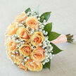 This arrangement reads like a love note in flowers: apricot-peach roses gathered into a perfect rounded cluster, their creamy petals overlapping like silk, surrounded by a halo of tiny white gypsophila that adds an airy, weightless quality. The stems are neatly bound in a length of peach satin ribbon studded with small pearls, a tactile detail that suggests careful, old-fashioned care. Large, glossy emerald leaves are interspersed throughout, giving the bouquet a lush, living frame that contrasts the soft pastel blooms and helps each rose stand out. Photographed in soft daylight, the palette is calm and refined, the neutral backdrop allowing the peach tones to glow with quiet radiance. The scent is fresh and understated - green-cut foliage notes underlining the delicate sweetness of rosés and the barely-there powder of baby's breath - a fragrance that suits weddings, vow renewals, or thoughtful anniversary gifts. As a florist serving Brent Cross and nearby Cricklewood and Golders Green, I imagine this as the perfect piece to carry down an aisle or to deliver for a small, heartfelt celebration; Brent Cross Flowers prepares bouquets like this with care, ensuring stems arrive fresh and comfortably arranged. The tactile ribbon, the layered petals, and the gentle light together tell a story of anticipation and calm joy, a keepsake of a day remembered for tenderness rather than spectacle.