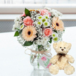 A serene, celebratory arrangement sits on a glossy white surface, its reflection faintly echoing the soft colours above. At the centre of the image is a compact pastel bouquet, gathered in a clear glass jar-style vase that reveals the neat stems within. The flowers are thoughtfully arranged: pale pink roses with layered, velvety petals mingle with soft peach gerbera daisies, whose dark chocolate-coloured centres draw the eye. Around them, clusters of white chrysanthemums with fresh lime-tinged centres provide structure and brightness, while delicate sprays of baby's breath float through the design like a fine mist, softening every gap. Touches of dark green foliage peek out here and there, grounding the arrangement and giving it a just-picked feel, as if it has come straight from a nearby garden rather than the bustling streets around Brent Cross. Wrapped around the neck of the vase is a piece of rustic natural twine, tied in a simple bow. Hanging from it is a small, light-coloured gift tag decorated with a subtle pink motif-perhaps a heart or flower-and faint green script, suggesting a handcrafted or specially selected gift ready to be delivered across the local area. To the right of the vase a classic cream teddy bear sits upright, its plush fur soft and slightly textured. The bear's dark bead eyes and brown stitched nose give it a gentle, reassuring expression, while a narrow dark brown ribbon is tied neatly around its neck. Behind them, the background is softly blurred and filled with bright natural light, allowing the arrangement and bear to remain the focus. Together they create an atmosphere of tenderness, ideal for anniversaries, birthdays, or simply sending a warm thinking of you from Brent Cross.