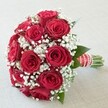 This vibrant bouquet, expertly arranged by a Brent Cross florist, holds center stage against a soft, neutral background that illuminates its striking colours and balanced form. Overflowing with fully bloomed, plush deep red roses, each flower showcases rich, velvety petals spiraling outward in layers of passion and beauty. Nestled between the dramatic reds are clusters of light, airy white baby's breath, giving the bouquet a fleeting sense of softness and depth, as if touched by the gentle breeze that moves through Brent Cross's neighbourhood streets. Subtle hints of dark green leaves peek through, grounding the bouquet in a fresh, natural palette. The flowers are gathered densely in a rounded, domed composition, with stems precisely bundled and wrapped in a luxurious red satin ribbon. Elegant white stripes and a cascade of small, shimmering pearls accent the handle, lending an extra note of refinement. Captured in soft, even daylight, every petal and detail is revealed-ready for wedding celebrations, anniversaries, or a meaningful local delivery to homes near Hendon or Finchley Road. This bouquet is more than a floral arrangement; it's a lasting expression of love, hope, and the artistry at the heart of Brent Cross Flowers.