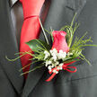 This close-up of a rose boutonnière showcases the refined detail and quiet romance that a local Brent Cross florist might prepare for a wedding morning. Dominating the design is a beautifully shaped hot pink rose, its spiralling petals close and plush, forming a tight, elegant rosette that feels both modern and classic. The colour leans toward a strong, saturated pink, making it stand out vividly against the dark suit it is designed to complement. Tucked around the rose, sprays of baby's breath-gypsophila-add a halo of tiny, papery white blooms, each stem branching into multiple little flowers that soften the overall look, suggesting delicacy and enduring love. Fine strands of decorative grass loop outward in gentle arcs, framing the rose without overwhelming it, like the graceful sweep of paths around nearby Brent Cross Park. Behind the main bloom, a single deep green leaf, smooth and slightly waxy, gives the boutonnière form and stability, its curved shape cradling the rose and catching bits of light along its surface. The stems are neatly wrapped at the base in a slim red ribbon, tied into a tidy, understated bow whose satin texture provides a subtle gleam and adds a celebratory note. Resting against a charcoal or black lapel, the boutonnière feels fresh and thoughtfully crafted, perfect for same-day delivery to homes and venues across Brent Cross. You can almost imagine the wearer adjusting it in a mirror before stepping out, the soft fragrance of rose and the airy feel of gypsophila accompanying them towards a ceremony filled with family, friends, and heartfelt moments.