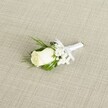 A single white rose boutonniere lies diagonally across a softly textured, linen-like beige fabric, captured as if resting on a groom's suit laid out before a Brent Cross wedding. The rosebud is creamy white with a hint of ivory warmth at the center, its velvety petals still mostly furled, suggesting a bloom cut at peak freshness that might have come straight from a local North London flower market. Around the rose, tiny white baby's breath blossoms create a delicate, cloud-like halo, each miniature flower adding lightness and air, like soft mist over the paths in nearby Brent Cross and Hendon Park. Slender, deep green leaves and a few feathery, fern-like sprigs cradle the central bloom, their glossy and matte textures contrasting gently and giving the boutonniere a quiet, natural depth. The stems are neatly wrapped in pristine white satin ribbon, the finish smooth and slightly reflective, tied into a small, careful bow that sits just below the flower head, ready to be pinned to a lapel for a ceremony in a local registry office or a nearby synagogue. The background fabric has a visible diagonal weave, subtly echoing the lines of a tailored suit, while the soft, diffused lighting adds a gentle glow to the rose, highlighting every curve of petal and the fine structure of the baby's breath. The image conveys calm anticipation and understated elegance, perfectly suited to illustrate groom or groomsmen accessories, classic wedding flowers, or formal event styling in and around Brent Cross, while conveying the tactile care of an experienced local florist who understands how important these tiny details feel on the day.