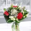 A contemporary mixed bouquet in a clear glass vase sits in soft window light, its colours and textures unfolding like a quiet celebration in a Brent Cross living room. At the heart of the arrangement, a single coral-red rose and a plush crimson carnation glow with intensity, their velvety petals layered and full, almost pulsing with emotion. Around them, clusters of delicate white chrysanthemums and tiny daisy-like flowers create a gentle halo, their small, crisp petals forming a cloud of softness that seems to float between the bolder blooms. Creamy white lilies, some just beginning to open and others still in neat green-tipped buds, rise gracefully through the composition, adding height and a sense of elegant anticipation, as if the bouquet will look slightly different and more open with each passing day. Wide, glossy emerald leaves are artfully folded and curved around the base, encircling the stems in sleek arcs that give the design a modern, sculpted silhouette, much like the stylish arrangements often seen in homes along Hendon Way or near the Brent Cross Shopping Centre. The glass vase catches and reflects the natural daylight, so that every leaf vein and petal edge appears sharp and luminous, enhancing the sense of freshness you might notice when this bouquet is delivered to a doorstep. Though the image is still, you can almost sense a light, green fragrance with a hint of sweetness, the sort of clean floral scent that quietly lifts the atmosphere of a room, perfect for a birthday, a small celebration, or simply bringing a sense of calm beauty to everyday life in NW11.