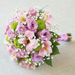 On a light grey, gently crumpled linen cloth, this bouquet is set down lengthwise as if resting between bridal portraits, its colours glowing softly against the neutral background. The arrangement is domed and compact, a hand-tied cluster of flowers chosen for their harmony and contrast. Dominating the eye are lush lavender roses, open to their full, rounded form, with petals that look almost touchably soft, swirling in graceful spirals from centre to edge. Nestled among them are clusters of pink and creamy-white alstroemeria, each bloom flaring outward with slender petals streaked with honey-yellow tones and delicate freckles, bringing a sense of movement and fine detail. Bright green button chrysanthemums stand proudly between the pastels, their small, globe-shaped blooms adding texture and a bold pop of chartreuse that immediately freshens the palette, much like the greenery in the landscaped spaces around Brent Cross Shopping Centre. Scattered wisps of baby's breath drift through the bouquet, their tiny, snow-like flowers forming a gentle haze that softens lines and adds an ethereal air. The stems are carefully aligned and bound with a wide stretch of pink satin ribbon, smooth and slightly glossy, forming a comfortable, polished handle. Twined around the ribbon, a single strand of small, pearl-like beads glints subtly, offering a refined bridal accent that feels both classic and modern. Below the ribbon, the cut stems extend neatly, revealing natural greens and browns that highlight the bouquet's fresh, garden-inspired origins. Designed by skilled local florists offering same-day delivery across Brent Cross, Hendon, and nearby areas, this bouquet suits wedding aisles, engagement celebrations, or any heartfelt occasion where soft romance and understated luxury are desired.