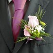 There's an uplifting elegance to this Brent Cross buttonhole, expertly arranged for wedding days and special events. The key visual note is a blush pink rose, its petals soft and open, exuding the gentle fragrance of a North London morning. Tucked alongside, a partially unfurled pink lily or alstroemeria-with signature fuchsia stripes-adds delicate detail, while a vibrant green chrysanthemum injects a fresh spark reminiscent of suburban parks. Airy gypsophila brings a touch of romance, weaving lightly through the blooms and enhancing the boutonniere's inviting texture. Wispy ferns and slender glossy foliage fan outward, adding fullness and natural grace, while a neat lavender satin ribbon is knotted at the base to accentuate the tie's vibrant magenta. The dark grey suit and colourful accessories create a harmonious finish, reflecting Brent Cross's lively yet refined spirit. This thoughtfully crafted piece is more than a flourish; it's a celebration in miniature, ideal for moments of unity, love, and joy that define milestones in the community.