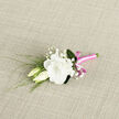 This carefully crafted boutonniere rests on a light, woven fabric that gives the scene a warm, tactile backdrop, as if prepared on a florist's worktable in Brent Cross moments before a celebration. The focal point is a fully opened white lisianthus, its petals plush and ruffled, layered like soft waves of cream. The bloom's shape is rounded and generous, with a gentle luminosity that suggests purity and quiet confidence. Tucked closely beside it are two lisianthus buds, slender and still wrapped in their outer petals, their tips showing subtle tones of ivory and fresh green. These buds mirror the idea of new chapters and unfolding stories, fitting for weddings or milestone events. Delicate sprays of baby's breath are threaded throughout, each tiny white blossom dotting the design with fine, cloud-like accents that add volume without weight. Slim strands of greenery and fine, grass-like leaves arc around the flowers, bringing in a touch of the garden and a sense of natural movement, much like the soft rustle of leaves you'd hear walking near Brent Cross parks. At the base, the stems are neatly aligned and wrapped in a bright pink satin ribbon, smooth and slightly glossy, catching the light with a modern sheen. A precise little bow completes the wrap, its playful loops giving a fresh, contemporary twist to the otherwise classic palette of white and green. The overall impression is one of refined tenderness and composed joy, making this boutonniere a perfect finishing touch for a groom, usher, or prom guest preparing for a memorable day in and around Brent Cross.