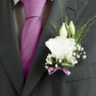 Pinned close to the heart on a charcoal suit jacket, this wedding boutonnière looks like a small, handcrafted tribute to love, carefully designed for a Brent Cross groom. The central feature is a pure white lisianthus bloom, its petals unfolding in relaxed, ruffled tiers that suggest both delicacy and resilience. The edges of the petals catch the light softly, giving the flower a gentle glow, while the inner folds remain slightly shadowed, adding depth and quiet drama. Just behind, two lisianthus buds lift their tapered heads, still wrapped in soft, green-tinged sepals, capturing that feeling of hopeful expectation that lingers before the vows are spoken at local venues around Brent Cross and Cricklewood. Around this central cluster, airy sprigs of baby's breath float like little clouds, their tiny round blossoms punctuating the design with delicate, white accents. Thin, linear stems of greenery weave between the flowers, introducing fresh, natural lines that soften the strong geometry of the suit lapel, almost as if a sliver of nearby Brent Reservoir's calm greenery had been gathered and woven in. A single dark green leaf curls gently at the base, shaped into a subtle arc that cradles the white bloom. Tied snugly around the stems is a soft mauve ribbon, its satin surface catching a faint sheen, forming a neat bow that hints at romance without overwhelming the piece. The boutonnière carries the suggestion of a light, clean floral fragrance, freshly prepared by local hands and possibly delivered the same day from Brent Cross Flowers. Its overall effect is refined yet warm, a graceful detail that quietly underscores the joy and significance of the wedding day.