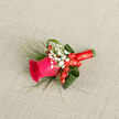 At the heart of this refined boutonniere sits a vivid fuchsia rose, a bloom so rich in colour it almost seems to glow against the neutral background beneath it. Each petal curves outward in velvety layers, the edges smooth and slightly ruffled, suggesting the rose has just opened to its most flattering point. Encircling the flower, delicate sprays of baby's breath add a cloud-like softness, their tiny white buds and blossoms forming a gentle halo that enhances, rather than competes with, the rose's intensity. Between these soft clusters lie bright red hypericum berries, glossy and plump, offering playful spots of colour that recall the cheerful atmosphere of celebrations around Brent Cross and Golders Green. The design is framed by deep green aspidistra leaves, skillfully curled and looped, their polished surfaces reflecting the light and leading the eye back to the central rose. Slim strands of grass stretch away from the arrangement in relaxed arcs, lending a sense of lightness and movement, similar to the swaying of trees along the nearby North Circular on a breezy day. The stem is wrapped with precision in a coordinating red satin ribbon, its texture smooth and sleek, giving a tailored, formal finish that would sit neatly on a groom's jacket or an evening suit. The boutonniere rests on a subtly textured, neutral fabric, which allows every colour-the fuchsia, white, green, and crimson-to stand out in clear, balanced harmony. It feels like a thoughtfully handcrafted piece, the kind of detail a local Brent Cross florist might prepare early in the morning for same-day delivery to a wedding party gathering in a nearby home.
