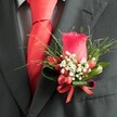 A detailed, close-up, high-angle photograph focuses on a refined boutonnière pinned neatly to the left lapel of a dark charcoal grey suit jacket, capturing the quiet elegance of formalwear on a special day in Brent Cross. At the heart of the arrangement, a single deep red rose stands proudly upright, its petals tightly furled into a perfect, velvety bud that suggests both romance and anticipation. Encircling the base of the rose, tiny clusters of white filler flowers, similar to baby's breath, create a soft halo of lightness, their star-like blooms gently breaking up the intensity of the red. Threaded among them are several small, smooth red berries, their glossy, oval forms mirroring the rose's rich hue and adding a subtle, tactile contrast. Behind the flowers, fresh greenery fans outward, with fine, grass-like blades radiating in slender arcs, supported by broader, darker green leaves that cradle and structure the design. A neatly tied red satin ribbon bow secures the stems, the sheen of the ribbon catching the light and seamlessly linking the berries and rose in one cohesive line of colour. Beneath the lapel, the knot of a bold red tie, likely silk or satin, sits perfectly centered, its vivid tone reinforcing the red accents of the boutonnière. The tie lies against a subtly patterned dress shirt, perhaps a fine grey-and-white check, adding a discreet layer of texture and sophistication. The overall mood is polished yet heartfelt, like a groom or prom guest stepping out near the Brent Cross Shopping Centre for photographs, every detail considered and composed for a memorable celebration.