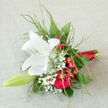 A graceful bridal-style bouquet is displayed as if ready to be lifted from the table and carried through a Brent Cross ceremony, its colours and textures telling a story of romance and joyful promise. At its heart is a single, striking white lily, open wide and immaculate, with creamy petals that radiate from the centre like soft, sculpted wings. The inner throat of the lily, touched with pale green and speckled pollen, draws the eye inward and hints at gentle fragrance. Surrounding this focal bloom are sumptuous dark red roses, their velvety petals layered so thickly that each flower looks plush and full, conveying depth of feeling and an enduring sense of commitment. Between the roses, clusters of rich red berries provide lively bursts of colour; their glossy skins catch the light, giving a subtle festive glimmer that would feel right at home at a winter wedding or anniversary gathering in a Brent Cross townhouse. Wisps of gypsophila drift throughout the bouquet, the tiny white blossoms forming clouds of softness that temper the bold reds and introduce a tender, almost whisper-like quality. Fine stems of airy greenery arc gently around the flowers, contributing movement, texture, and a natural, garden-inspired frame that keeps the design from feeling rigid. The stems are drawn together and wrapped in a slender ribbon of red satin, sleek and understated, forming a comfortable handle. The overall impression is one of poised elegance with a personal touch, suitable for walking down the aisle, renewing vows, or sending a meaningful you matter message to someone local. This bouquet reflects the balance of modern life and timeless sentiment that characterises Brent Cross, harmonising fragrance, colour, and texture into one considered gesture.