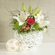The photograph depicts a stylish wedding-style table arrangement placed on a spotless white surface, set before a subtly textured beige backdrop that lets the flowers shine without distraction. At centre stage is a distinctive spherical vase in bright white, created from interwoven strands that form a hollow basket-like structure. Its open-weave design gives a modern, airy impression, perfectly complementing the light-filled interiors often found in Brent Cross event spaces and nearby Hendon function rooms. From this sculptural vase rises a full and balanced bouquet featuring elegant white lilies, romantic red roses, vibrant green pompon blooms, and an abundance of lush greenery. Several lilies are already in generous bloom, their wide, creamy petals curving gracefully backwards to expose golden stamens dusted with pollen and subtle, speckled throats. Slender green lily buds stretch above them, adding height and suggesting blossoms still to unfold. Tucked between the lilies, velvety red roses bring a sense of passion and intimacy; one especially prominent rose sits near the centre, its tightly layered petals forming a perfect, eye-catching spiral. Rounded, bright green dianthus or chrysanthemum pompons are nestled among the blooms, offering a playful, cushiony texture that contrasts with the smooth petals and adds a fresh, garden-like feel. Fine sprays of white gypsophila thread through the arrangement, softening edges and lending a delicate, cloud-like effect. Clusters of small red berries peek out from between the leaves, echoing the rose tones and introducing a subtle hint of seasonal festivity. Wispy asparagus fern fronds and darker, broader foliage spill over the rim of the vase, giving the design natural movement and depth. Positioned to the left of the main bouquet, two pale woven spheres quietly mirror the vase's texture, completing an elegant and harmonious centrepiece ideal for celebrations and wedding receptions around Brent Cross.