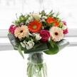 A lively and inviting bouquet bursts with dynamic colour and texture, expertly gathered by a local Brent Cross florist. The heart of the design glows with peach gerberas, their creamy petals softly transitioning into the brighter orange blooms that radiate joy-much like a sunbeam through the windows of a Brent Cross home. Magenta roses, rich and velvety, spiral throughout, infusing the bouquet with romance and elegance. Clusters of pale ivory alstroemeria and tiny white spray roses surface between the greens, their gentle perfume suggesting the crispness of a spring morning near Hendon Way. Encircling the flowers, broad aspidistra leaves fold gently, supplying structure and a designer's touch reminiscent of the curated gardens throughout Brent Cross. Additional luscious greenery spills naturally, providing fullness and contrast. Housed in a tall, simple glass vase, the fresh green stems are visible below, anchoring the arrangement with a sense of vitality. The soft, natural lighting highlights every shade and shape against a subtle background, keeping the focus on the beauty of the blooms. Overflowing with warmth and gratitude, this bouquet is perfect for lifting someone's spirits, marking an accomplishment, or adding a splash of seasonal colour to gatherings throughout Brent Cross.
