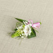 On a neutral oatmeal-toned fabric that looks like fine linen, this boutonniere glows softly, its white and green palette made warmer by the gentle lighting. The focal point is a single, open alstroemeria-like bloom in pure white, its petals rounded yet delicately tapered, decorated on the inside with subtle reddish-brown streaks that give it character without interrupting its calm elegance. Around it, smaller white buds cluster in close, echoing the forms of campanula or lily-of-the-valley and enhancing the sense of intimate detail you might expect from a Brent Cross Flowers design. Tiny specks of gypsophila are scattered throughout, like fine lace, introducing a dreamy, ethereal layer that makes the composition feel almost weightless. To the left, a vibrant, cone-shaped cluster of smooth green buds-resembling ornithogalum-adds a bold vertical accent and a modern, sculptural edge. Behind and beneath, glossy, dark green leaves form a stable, natural frame, while strands of narrow bear grass arc over and around the flowers, their lines reminiscent of the gentle curves of local pathways and garden borders. The stems meet neatly at the base, bound securely in a bright pink satin ribbon tied into a small, polished bow, the ribbon's sheen catching the light and signalling an occasion of joy: perhaps a wedding at a nearby hotel, a formal event, or a significant celebration with family and friends. The boutonniere feels both refined and approachable, as though created for someone who values subtle beauty and carefully considered details, ready to be pinned to a lapel and carried through an important day around Brent Cross.
