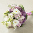 A carefully crafted bouquet is shown resting sideways on a pale beige, lightly textured fabric that might be linen or burlap, its understated surface allowing every bloom to stand out. Dominating the arrangement are full, velvety white lisianthus flowers, some wide open and revealing subtle yellow-green centres, others just beginning to unfurl. Their gentle curves suggest softness and intimacy, like a quiet smile. Nestled closely among them are clusters of stock in shades of soft pink and muted mauve, each stem covered in small, ruffled florets that build a romantic, almost cloud-like volume through the middle of the bouquet. Punctuating these pastels, spiky heads of purple allium or similar filler flowers emerge, their star-shaped florets creating a delicate, airy halo of lavender-purple that breaks up the smooth petal surfaces with lively texture. Fine, deep green blades of foliage loop and weave through the design, especially noticeable as they arc along the right-hand side, giving the bouquet a graceful sense of movement, as if a breeze had just passed through. The gathered stems are firmly bound with a rich fuchsia satin ribbon, its sheen enhanced by a narrow strand of tiny, round white pearls that run along the handle, suggesting classic bridal jewellery. The overall impression is one of refined romance, ideal for a bride walking down the aisle in Brent Cross or celebrating in a local venue tucked off the North Circular. The flowers appear fresh and full of life, conveying that quiet, meaningful joy that often accompanies weddings and intimate celebrations across this part of North West London.