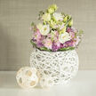 Sunlight seems to linger in the folds of creamy lisianthus as this round, lush bouquet breathes quietly on a bright white shelf. The dominant lisianthus-white and warm cream tones with a whisper of buttery yellow-forms soft, cloud-like blooms that are balanced by slender stems of blush-pink stock and darting clusters of purple allium that add architectural punctuation. Fresh green foliage fans through the arrangement, and a band of deeper green leaves neatly wraps the base, giving the bouquet a deliberate, tailored finish. The vessel is as much a statement as the flowers: a white, spherical vase formed from many interwoven, slender branches that create a lattice, revealing stems and water inside while feeling sculptural and hand-crafted. Beside it, two woven rattan orbs in natural, lightwood tones sit to the left, their crisscross construction mirroring the vase and introducing a soothing organic rhythm. The entire composition rests on a reflective white surface beneath a soft, greige textured wall-think linen wallpaper-so the pastels pop and the textures read clearly even at a distance. I imagine this piece arriving from Brent Cross Flowers for a bedside table in Childs Hill or as a calming centrepiece for a small gathering in Cricklewood; it reads as both contemporary decor and an intimate, fragrant gesture. The palette, scent, and tactility together create a welcoming, restorative presence that quietly lifts a room without demanding attention.