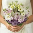 In a sun-dappled room near Brent Cross, an exquisite bridal bouquet is gathered gently in a bride's hands, the composition radiating effortless beauty and hope. The creamy whites of Lisianthus flowers unfurl in delicate ruffles, offset by pale, unopened buds that suggest anticipation and new beginnings. Around them, airy clouds of lavender and blush-pink stock bring a tender sweetness, their soft petals releasing a subtle perfume that calls to mind the spring blooms of North London parks. A distinctive base of vibrant purple Allium flowers forms a modern, star-like collar, adding depth and playful contrast to the bouquet's rounded, romantic shape. Arched loops of glossy bear grass wrap the midsection, their deep greens providing an elegant contour that reflects the flourish of Brent Cross's established avenues. The bride's carefully polished nails and the misty glimpse of an ivory lace gown in the soft background frame the flowers with quiet intimacy. This bouquet, dreamt up for a modern Brent Cross wedding, balances tradition and innovation, filling the air with gentle exuberance, and promising couples a natural sense of celebration and joy on their most unforgettable day.