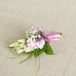 A delicately crafted floral corsage or boutonniere rests on a softly textured, oatmeal-toned linen background, the neutral fabric making every petal and ribbon detail stand out with quiet elegance. At the centre, a compact cluster of soft blush-pink blooms, reminiscent of miniature stocks or ruffled garden flowers, forms a gentle cushion of colour, their multi-layered petals showing subtle gradients from pale shell pink at the edges to a slightly deeper tone in the folds. Threaded between these blooms are airy sprigs of white baby's breath, each tiny floret like a floating cloud that adds lightness and intricate texture. Slight touches of deeper lavender or dusky purple flowers peep through, introducing a refined contrast without disturbing the overall pastel mood. On one side, several creamy white to pale green buds, similar to slender freesias, extend gracefully outward, closed and full of promise, suggesting flowers that will open later in the day. Broad, glossy green leaves cradle the arrangement, while fine wisps of fern-like foliage and grass-textured sprigs reach delicately beyond the main cluster, adding movement and a natural, garden-gathered feel. The stems are neatly bound and finished with a vivid fuchsia satin ribbon, tied into a precise, symmetrical bow whose smooth sheen catches the light, its short tails lying neatly across the fabric. The composition is evenly lit, revealing each vein, fold, and curve with clarity, and the overall mood is one of poised celebration-a refined floral accent perfect for a Brent Cross wedding, school prom, or graduation photo taken after a ceremony in nearby local parks or venues, where subtle, handcrafted details quietly elevate the day.
