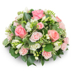 A softly domed, cushion-like floral tribute composed of tightly arranged soft pink roses and clusters of pristine white freesia, set against a clean white background so every delicate detail can be seen. The roses range from fully opened, velvety blooms with gently unfurling petals in shades from blush to warm salmon, to smaller spray roses that add texture and depth; each petal appears fresh and slightly dewy under soft, natural light. Scattered throughout the rounded form are pure white freesia sprays - some bells fully open, their slender throats revealing pale yellow stamens, others still in elongated buds that promise a later unfurling and a subtle, sweet fragrance. The flowers sit above a lush bed of varied greenery: broad, glossy dark green leaves that cradle the composition (think ruscus or salal) and finer variegated foliage edged in cream that brightens the overall palette, likely pittosporum or euonymus. The arrangement is dense and formal, hiding stems entirely to preserve a polished, comforting silhouette suited to a memorial or quiet celebration of life. Gentle shadows on the white surface give the piece three-dimensional presence without distracting from its serenity. As a local florist working from Brent Cross, I picture this piece arriving with respectful care to a chapel near Brent Cross Shopping Centre or gently placed at a family home off Hendon Way - a refined, fragrant expression of sympathy and quiet affection that reads as both contemporary and timeless.